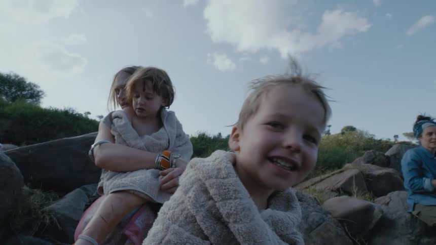 A woman holds her daughter as the girl and her brother sit wrapped in towels on large rocks outside