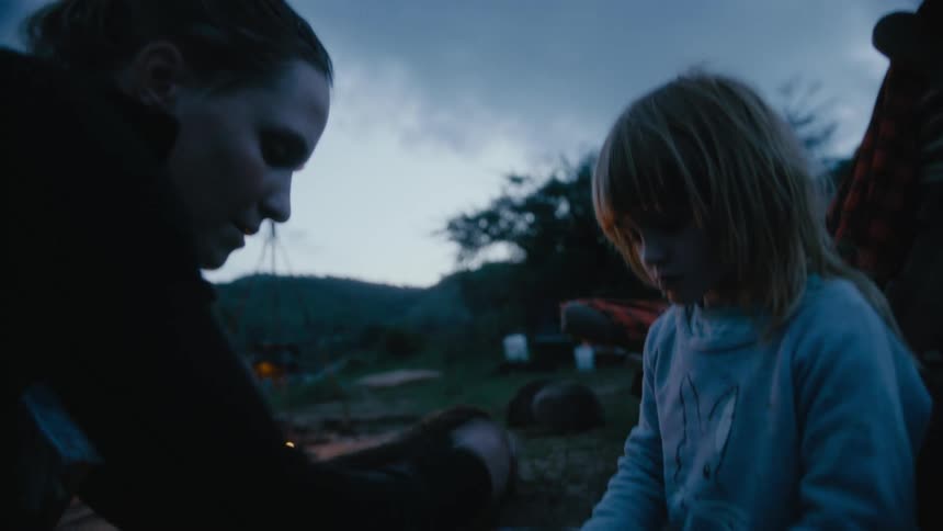 A young girl helps her mother cut food as a campfire burns beside them