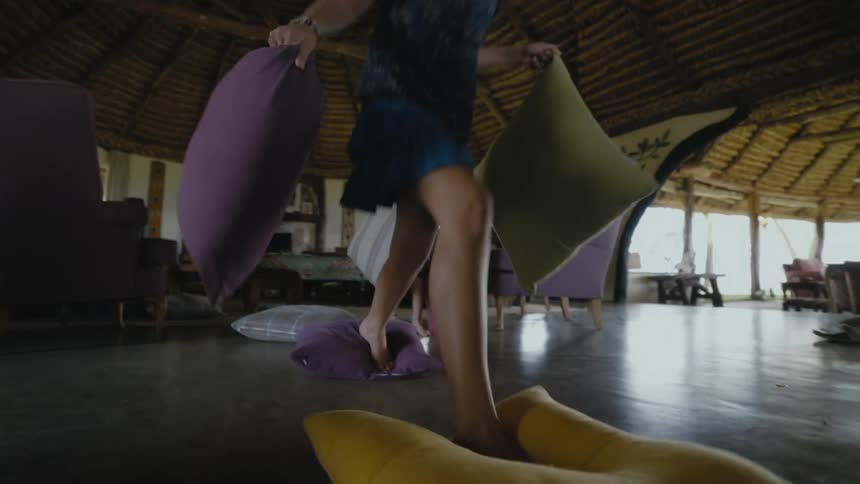 A young girl puts pillows on the living room floor and walks across them and her brother does the same behind her