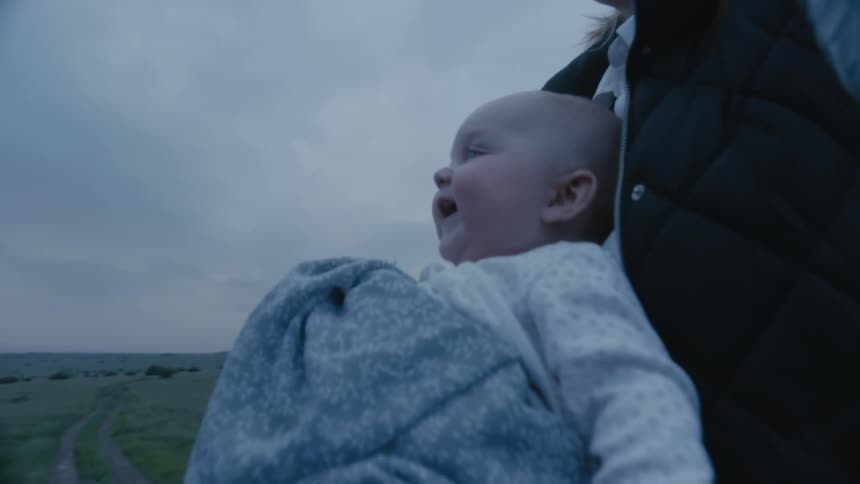 Older man makes faces at baby being held against blonde mother as dirt road leads toward field horizon in the background