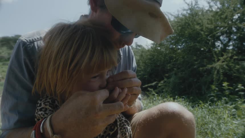 A man on a ranch holds his daughter in his lap and bring her fingers up to her nose to smell