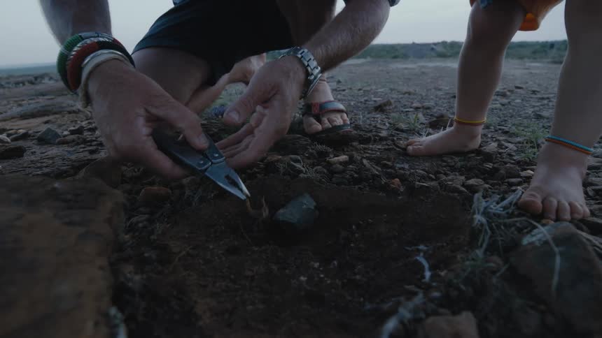 A man uses pliers to pick a scorpion up out of a hole while his barefoot toddler watches