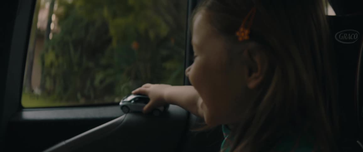 A little girl is sitting in a car that is traveling on a neighborhood street and she is smiling and playing with a toy car