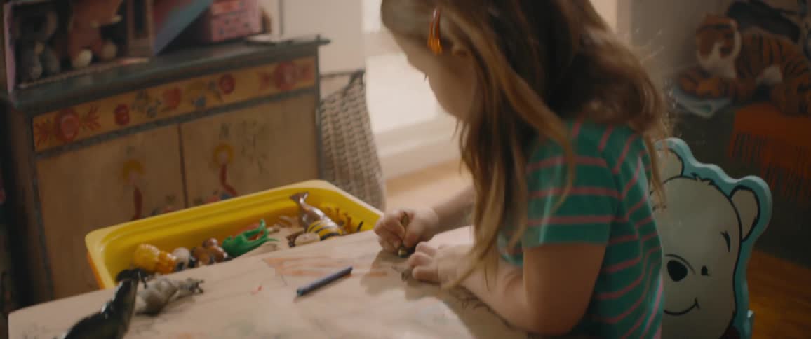 A girl uses a crayon to color at a small table covered in toys during the day