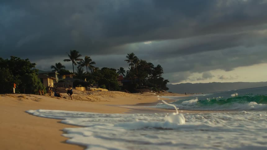 Waves roll over the sand on a beach as people stand watching near a beach house as dark clouds move overhead
