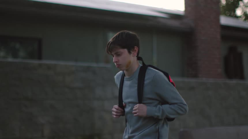 A boy wearing a backpack and holding the straps is walking by houses and trees