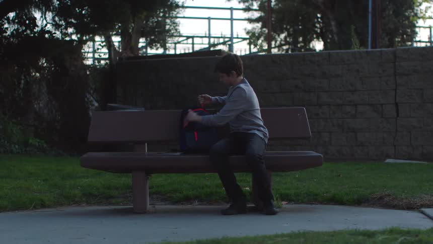 Teenaged boy sits on a park bench and aggressively removes a sheet of paper from his backpack