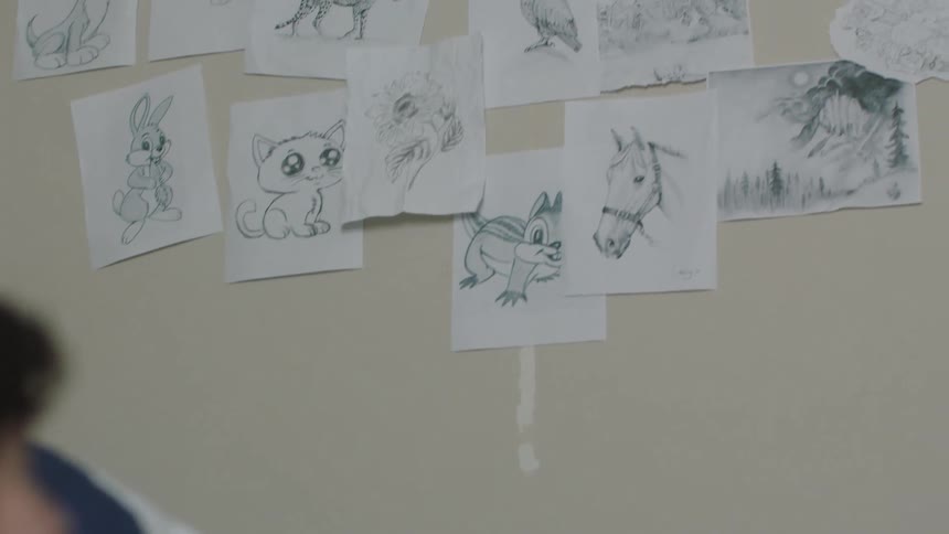 A boy is sitting at a table writing and he looks up at drawings on the wall