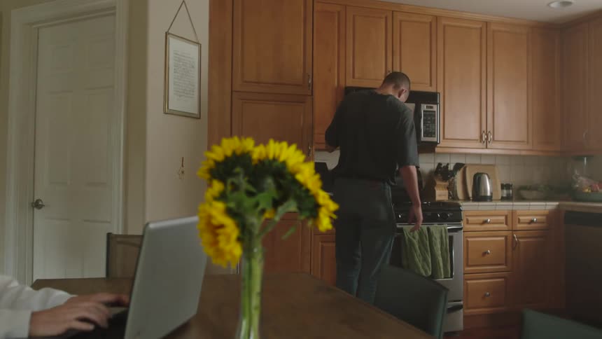 Woman sits at a kitchen table and types on a laptop while her husband walks by talking on his cellphone