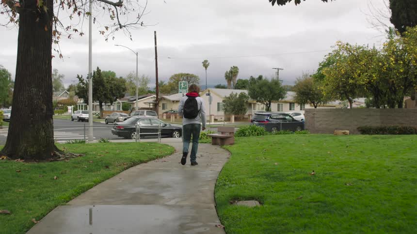 A boy with a backpack is sitting on a bench and he gets up and walks down the sidewalk to a black car parked on the street
