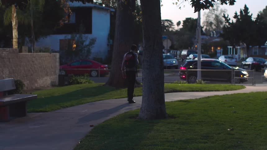Teenaged boy carrying a backpack walks through the park towards the street where many cars are parked