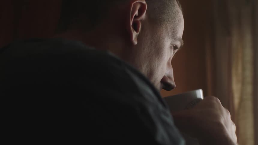 A man is standing in a kitchen holding a coffee mug and he takes a drink