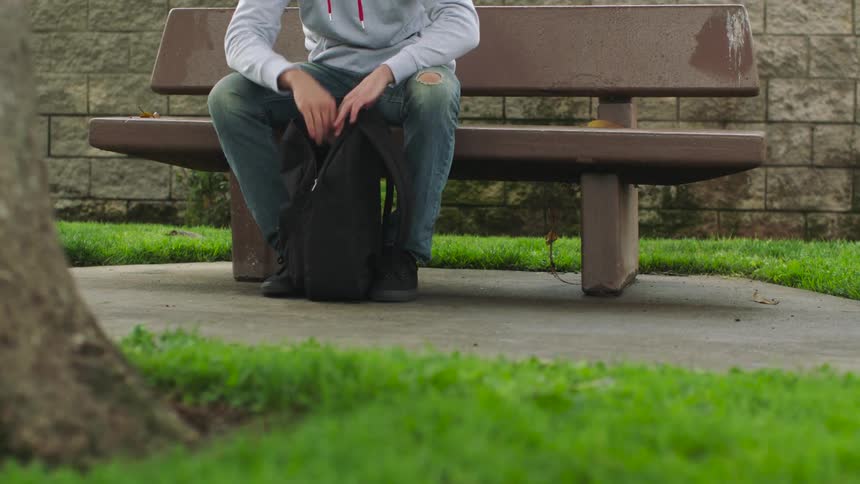 Woman with gray sweater and ripped jeans sits on bench and opens black backpack in park