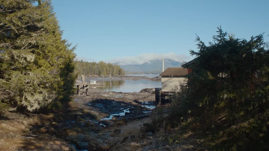 The sun shines on a forest lake with a cabin and mountains in the distance under a clear blue sky