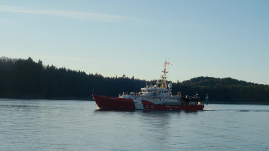 A ship travels a waterway near a forested coastline