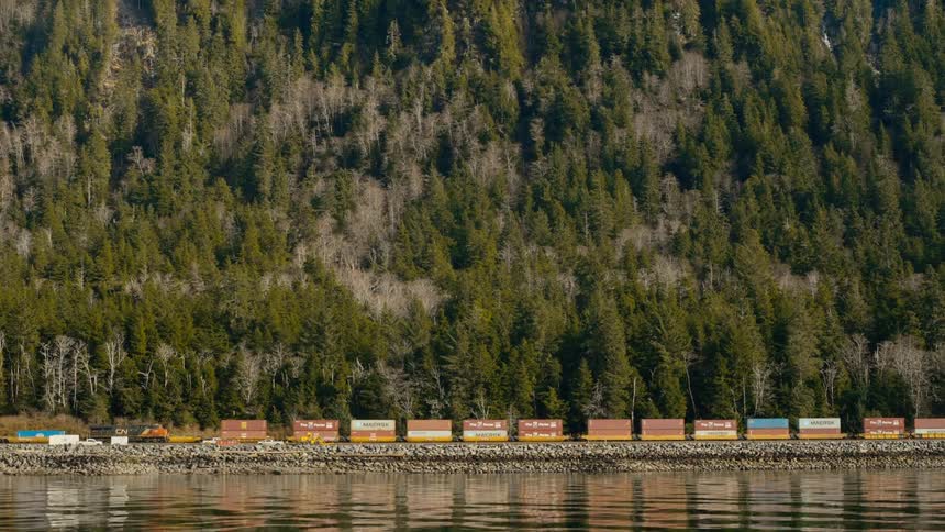 A train pulls cargo alongside a lake and forested mountains