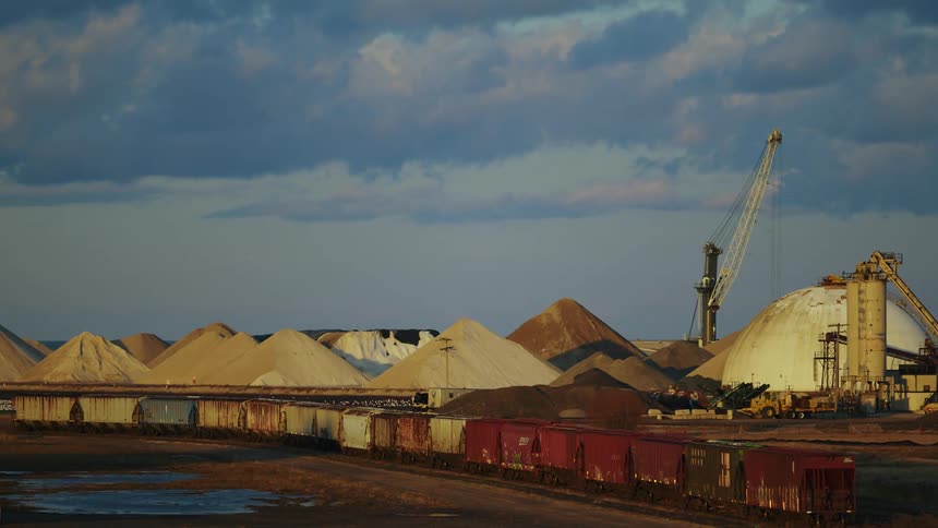 A train is near a piles of stone on a loading dock and a crane is high in a cloudy sky