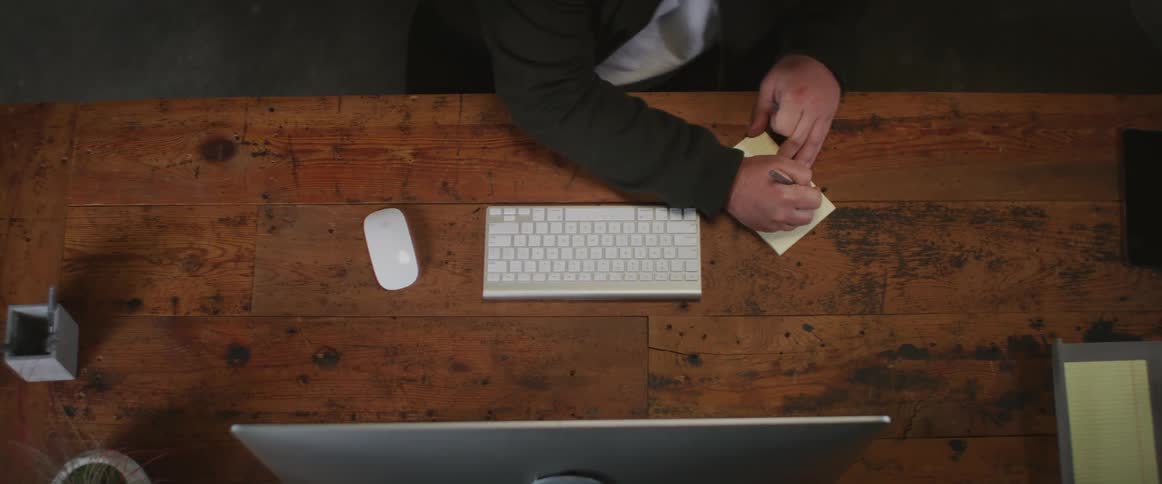 Man with green jacket and white shirt writes down in small notebook sitting at desk in front of computer