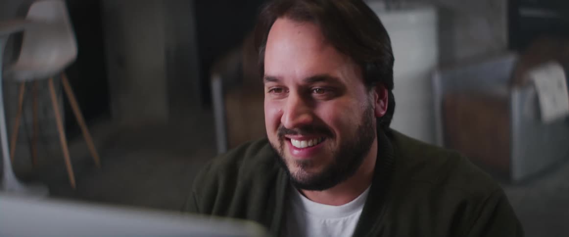 A man is smiling as he sits at a desk typing on a keyboard and looking at the computer monitor