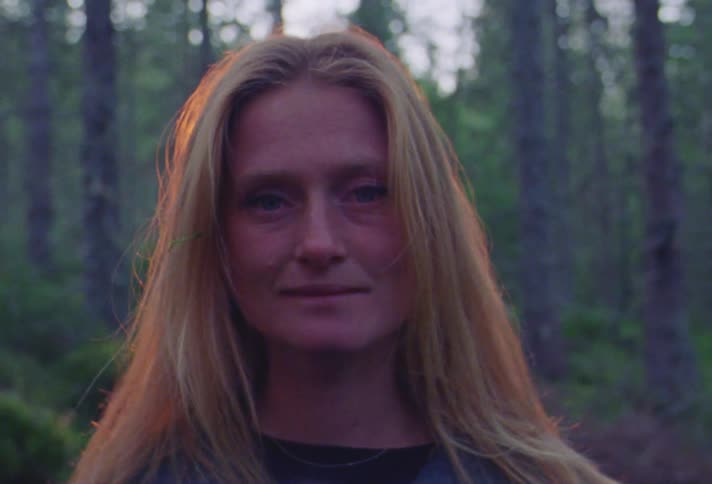 A woman standing in a forest looks into the camera while the light shining on her face fades