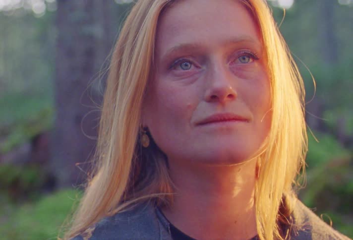 A woman sits up in the woods looking disoriented but pleased