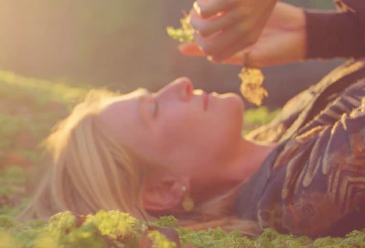 A woman resting on the ground picks up and examines a plant before rising out of frame