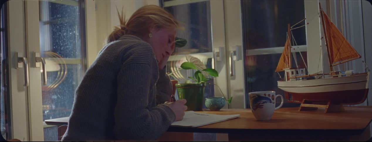 A woman is sitting at a table writing in a notebook and she tears out a page and throws it away