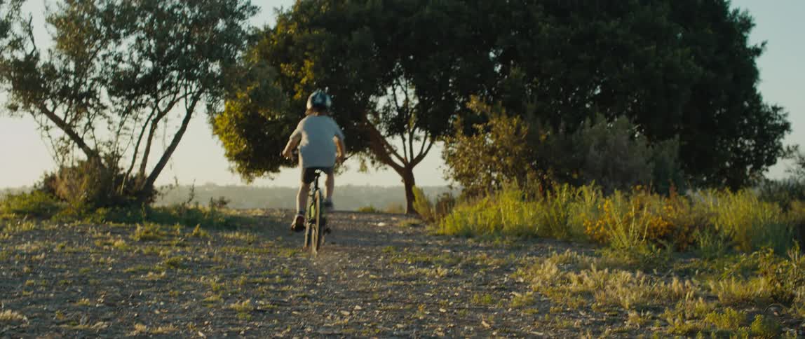A child rides a bicycle along a dirt path through a field during the day