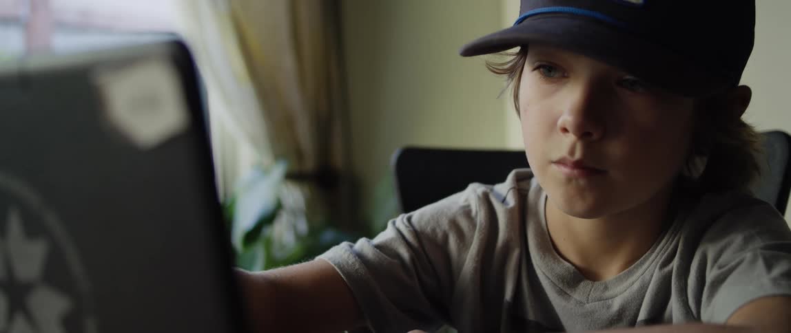 Kid wearing baseball cap sits at table making selections on laptop touchscreen