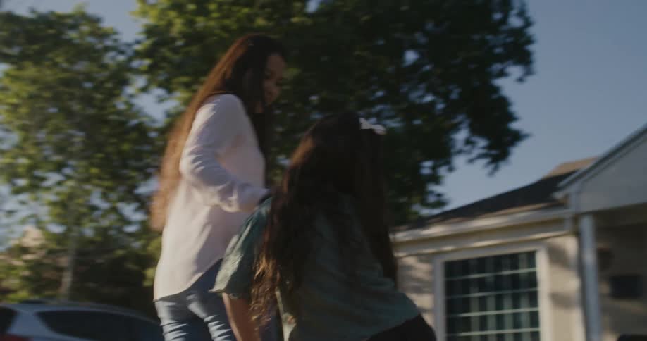 A woman with long dark hair is pulling a little girl in a red wagon and they stop and the girl gets out and they walk toward a house