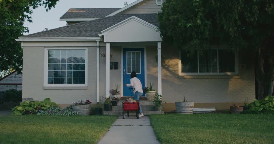 A woman pulling a wagon behind her walks onto the porch of a house and knocks on the door