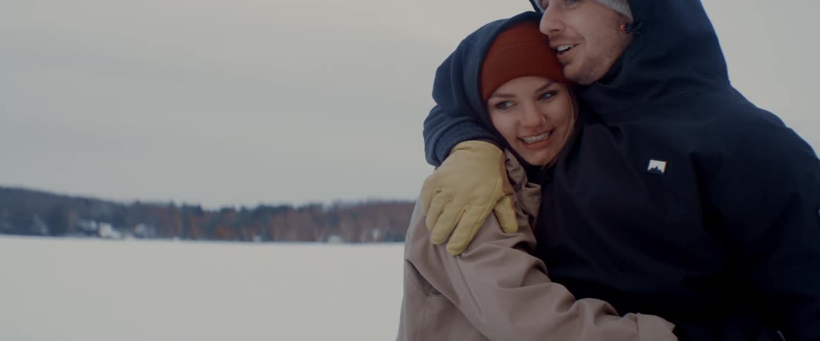 A man hugs a woman outside on a winter day with a forest of trees in the distance