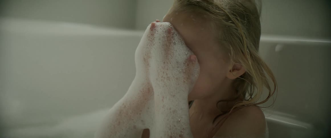 A little girl in a tub scoops up bubbles and covers her face laughing and smiling