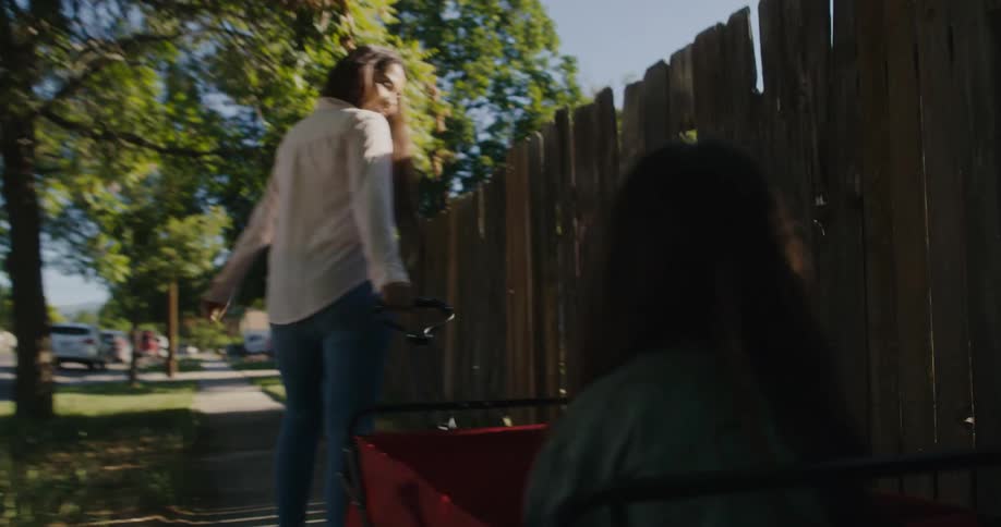 A woman is pulling a red wagon with a little girl in it and walking down a neighborhood sidewalk