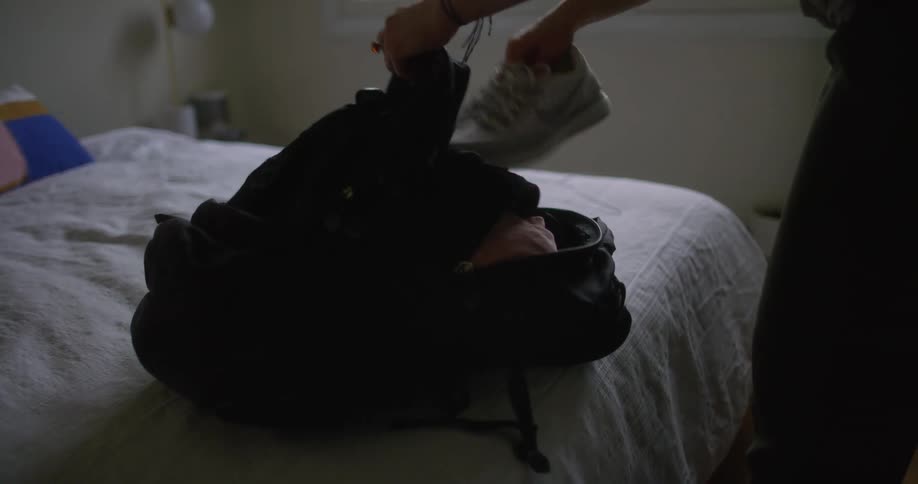A woman puts clothes into her backpack in her bedroom before leaving with it