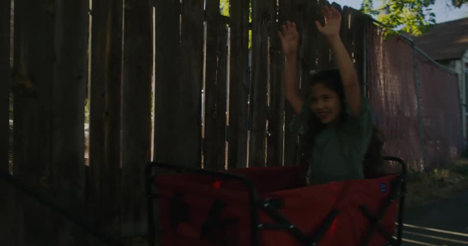 A little girl riding in a red wagon raises her arms in joy as mom pulls her down the sidewalk