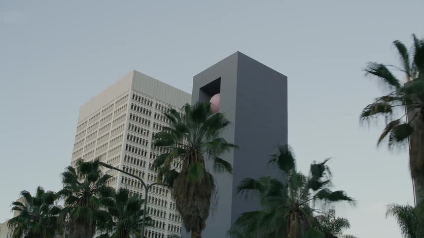 A sculpture of a tall tower with a pink ball inside stands among skyscrapers downtown