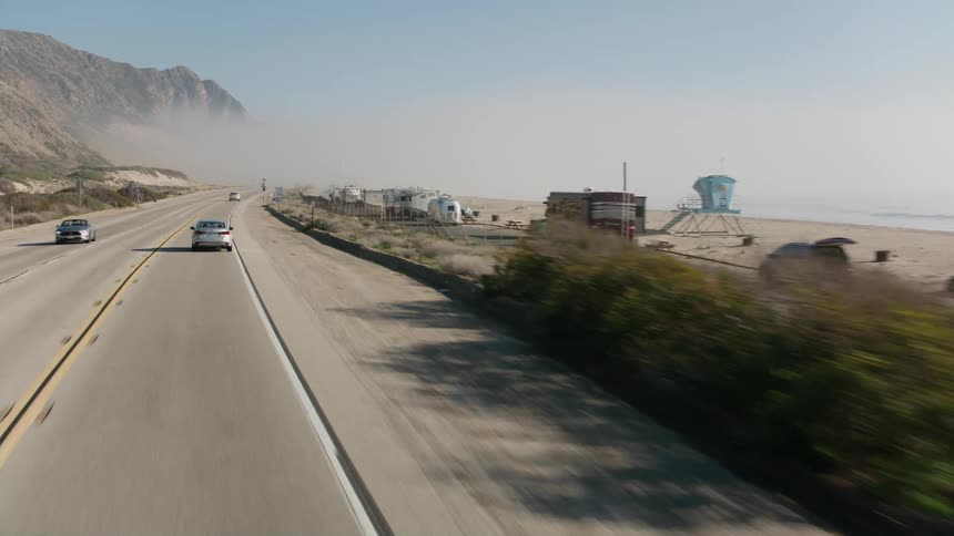 Vehicles travel an oceanside highway with campers parked on the beach