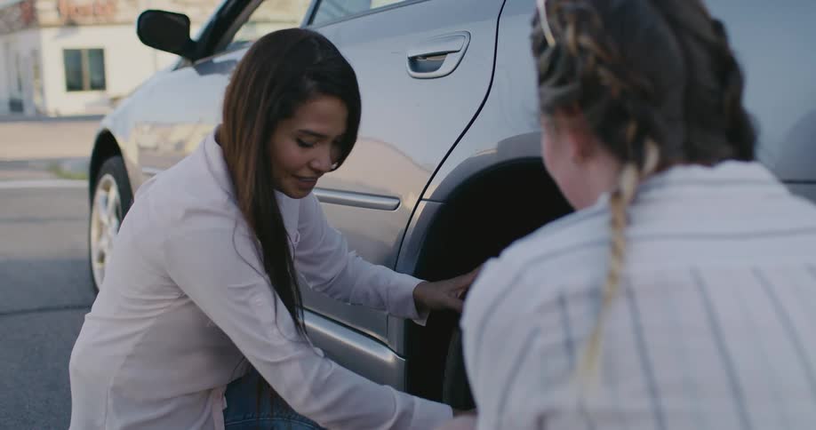 A woman talks and smiles as she changes a tire for another woman