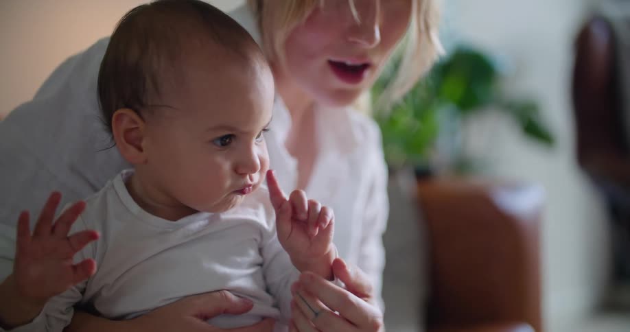A woman sitting on a sofa holds a baby in her lap as it reaches for something out of frame