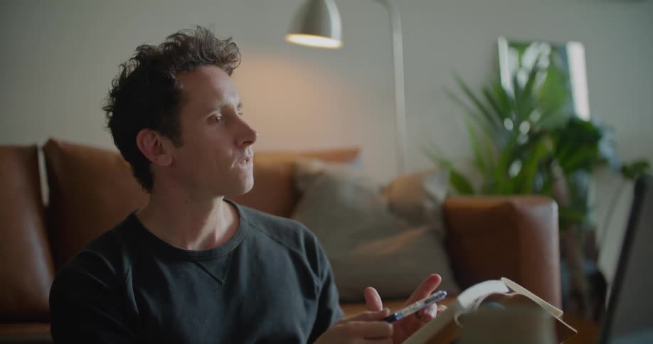 A man sits at home and snaps his fingers and taps his pen on a notebook as he talks with someone on a laptop