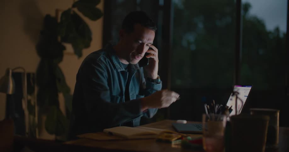 A man is talking on the phone and looking at a laptop at night