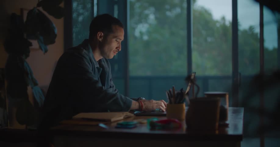Man with blue shirt eats food and works on laptop sitting at table at dusk