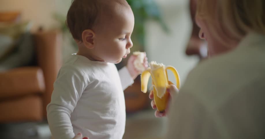 Mother holds up a peeled banana and her baby takes a bite then laughs