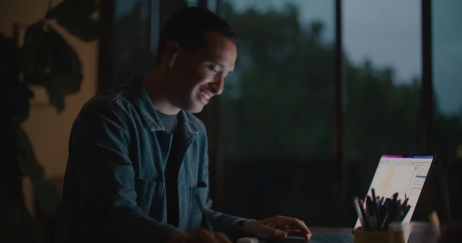 A man is typing on a laptop and sitting indoors at night
