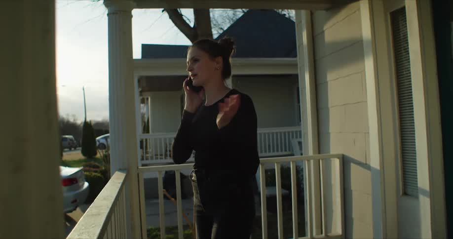 A woman talks on her smartphone while standing outdoors on the porch of a house during the day