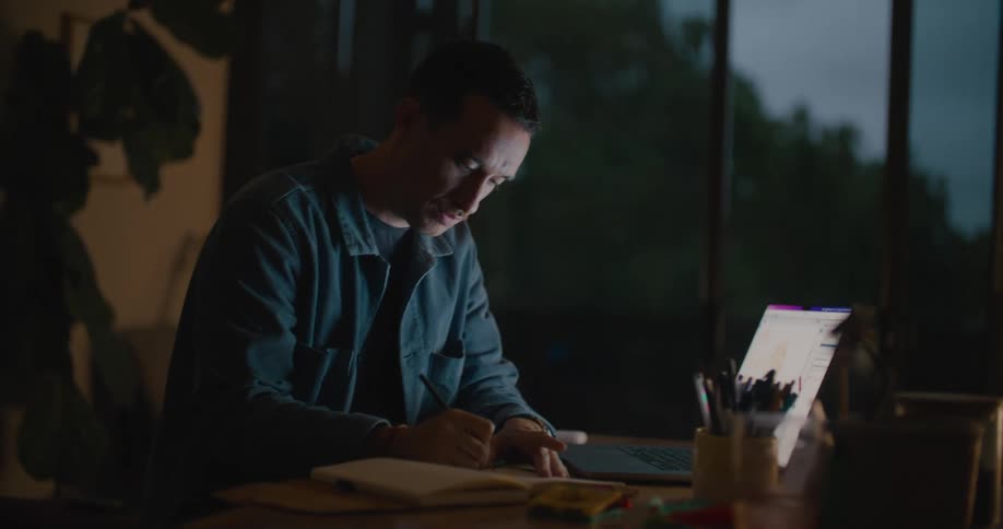 Man sitting at the desk using his laptop while writing something on his notebook