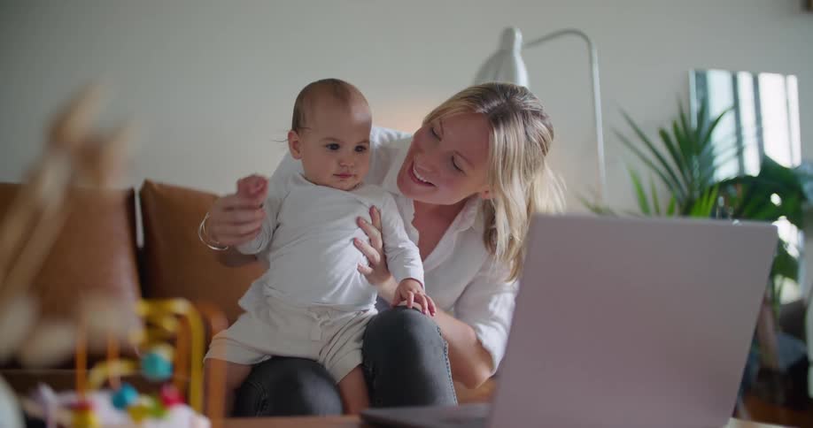 Blonde mother with shirt and pants holds baby sitting on brown couch waving at person on laptop in living room at home