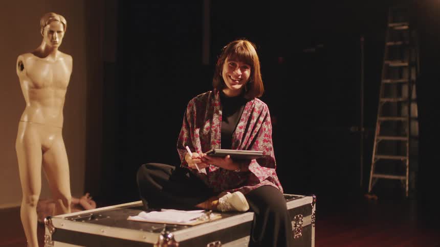 A woman holding a graphics tablet while sitting atop a large trunk smiles into the camera before turning her attention to her tablet