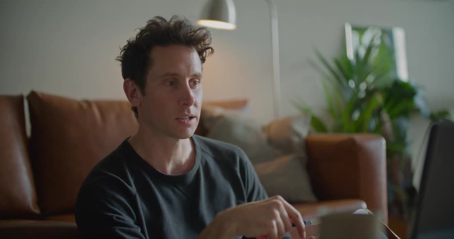A man sits on the floor in front of a couch and talks to someone on his laptop